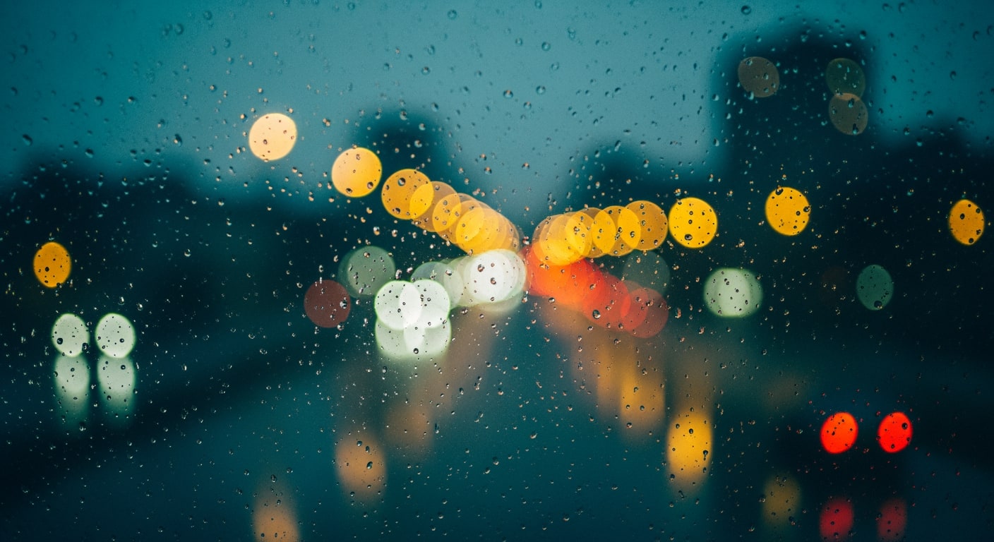 A rain-streaked window pane with blurred city lights in the distance at night, creating a soft glow.